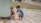 Two children are sitting on a checkered pavement near a large container. One child is filling a small canister with water from a makeshift tap while the other drinks from their hands. The background shows people and discarded plastic items, suggesting a sense of resourcefulness and adaptation.