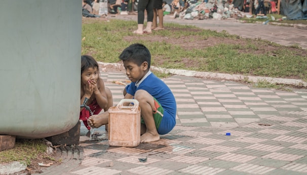 Two children are sitting on a checkered pavement near a large container. One child is filling a small canister with water from a makeshift tap while the other drinks from their hands. The background shows people and discarded plastic items, suggesting a sense of resourcefulness and adaptation.