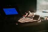 A close-up view of a music production device featuring a blue-lit screen displaying text and various buttons and controls. The equipment is dimly lit, creating a sense of focus and detail on its interface.