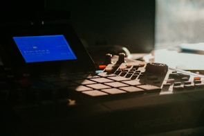 A close-up view of a music production device featuring a blue-lit screen displaying text and various buttons and controls. The equipment is dimly lit, creating a sense of focus and detail on its interface.
