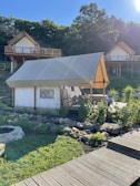 A glamping tent is set in a lush, green environment with wooden cabins on stilts in the background. The tent is surrounded by a garden with rocks and plants, and a wooden walkway leads up to it. The area is bathed in sunlight, casting shadows and creating a peaceful atmosphere.