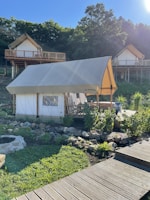 A glamping tent is set in a lush, green environment with wooden cabins on stilts in the background. The tent is surrounded by a garden with rocks and plants, and a wooden walkway leads up to it. The area is bathed in sunlight, casting shadows and creating a peaceful atmosphere.