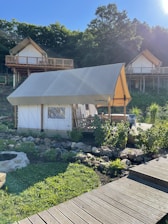 A glamping tent is set in a lush, green environment with wooden cabins on stilts in the background. The tent is surrounded by a garden with rocks and plants, and a wooden walkway leads up to it. The area is bathed in sunlight, casting shadows and creating a peaceful atmosphere.