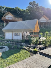 A glamping tent is set in a lush, green environment with wooden cabins on stilts in the background. The tent is surrounded by a garden with rocks and plants, and a wooden walkway leads up to it. The area is bathed in sunlight, casting shadows and creating a peaceful atmosphere.