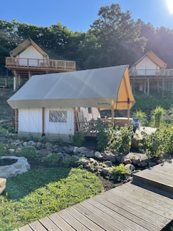 A glamping tent is set in a lush, green environment with wooden cabins on stilts in the background. The tent is surrounded by a garden with rocks and plants, and a wooden walkway leads up to it. The area is bathed in sunlight, casting shadows and creating a peaceful atmosphere.