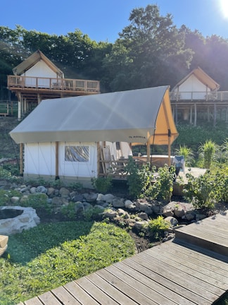 A glamping tent is set in a lush, green environment with wooden cabins on stilts in the background. The tent is surrounded by a garden with rocks and plants, and a wooden walkway leads up to it. The area is bathed in sunlight, casting shadows and creating a peaceful atmosphere.