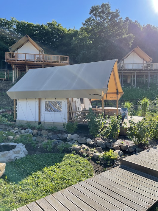 A glamping tent is set in a lush, green environment with wooden cabins on stilts in the background. The tent is surrounded by a garden with rocks and plants, and a wooden walkway leads up to it. The area is bathed in sunlight, casting shadows and creating a peaceful atmosphere.