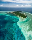 An aerial view of a tropical island surrounded by vibrant turquoise waters and coral reefs. The island is lush with greenery and several buildings are visible near the shoreline. The sky is partially cloudy, adding to the serene and picturesque scene.