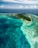 An aerial view of a tropical island surrounded by vibrant turquoise waters and coral reefs. The island is lush with greenery and several buildings are visible near the shoreline. The sky is partially cloudy, adding to the serene and picturesque scene.