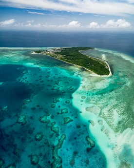An aerial view of a tropical island surrounded by vibrant turquoise waters and coral reefs. The island is lush with greenery and several buildings are visible near the shoreline. The sky is partially cloudy, adding to the serene and picturesque scene.