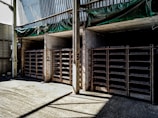 The image shows a warehouse or storage area with three large shelving units filled with rectangular items. The shelves are numbered 1, 2, and 3. The walls and ceiling are made of concrete and metal, and there is a green tarp hanging above the shelves. The area is lit by natural light coming from an opening on the left side.