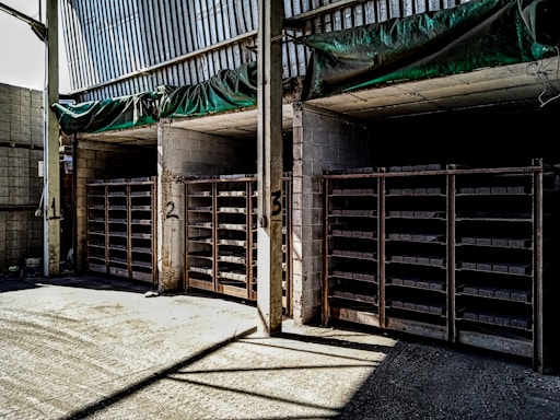 The image shows a warehouse or storage area with three large shelving units filled with rectangular items. The shelves are numbered 1, 2, and 3. The walls and ceiling are made of concrete and metal, and there is a green tarp hanging above the shelves. The area is lit by natural light coming from an opening on the left side.