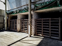 The image shows a warehouse or storage area with three large shelving units filled with rectangular items. The shelves are numbered 1, 2, and 3. The walls and ceiling are made of concrete and metal, and there is a green tarp hanging above the shelves. The area is lit by natural light coming from an opening on the left side.