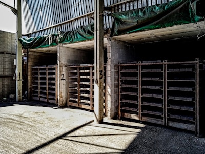 The image shows a warehouse or storage area with three large shelving units filled with rectangular items. The shelves are numbered 1, 2, and 3. The walls and ceiling are made of concrete and metal, and there is a green tarp hanging above the shelves. The area is lit by natural light coming from an opening on the left side.