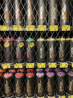 A neatly organized display of black spray paint cans is behind a wire fence. Each can is labeled with the word 'BLACK' in large text, and they are aligned in rows on shelves. Energy and vibrancy are added by colorful caps in shades of red, purple, and yellow. Price tags are visible below each row.