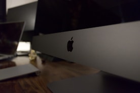 A sleek, modern workspace featuring a large computer monitor with the Apple logo prominently displayed. The setting includes a wooden desk surface with another laptop visible in the background, suggesting a technology-focused environment.