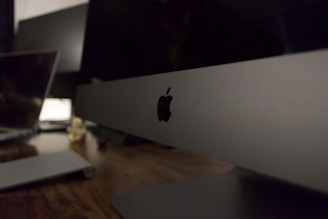 A sleek, modern workspace featuring a large computer monitor with the Apple logo prominently displayed. The setting includes a wooden desk surface with another laptop visible in the background, suggesting a technology-focused environment.