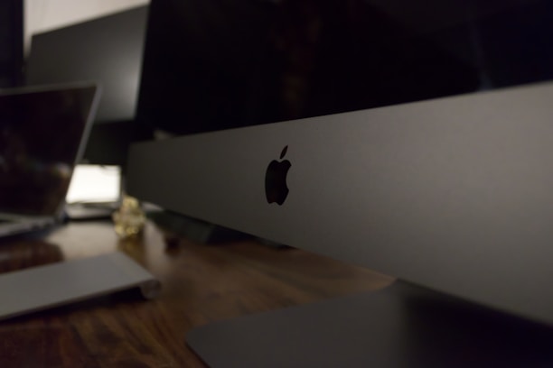 A sleek, modern workspace featuring a large computer monitor with the Apple logo prominently displayed. The setting includes a wooden desk surface with another laptop visible in the background, suggesting a technology-focused environment.