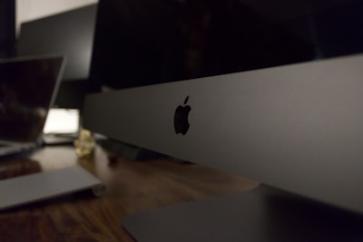 A sleek, modern workspace featuring a large computer monitor with the Apple logo prominently displayed. The setting includes a wooden desk surface with another laptop visible in the background, suggesting a technology-focused environment.