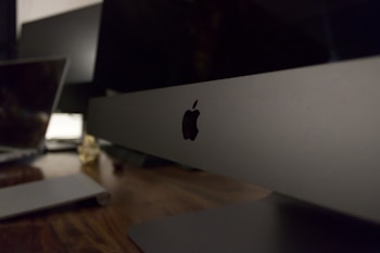 A sleek, modern workspace featuring a large computer monitor with the Apple logo prominently displayed. The setting includes a wooden desk surface with another laptop visible in the background, suggesting a technology-focused environment.