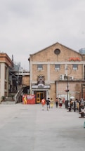 An outdoor space featuring a large brick building with industrial architectural elements. There are several people walking in the open area, creating a sense of casual movement. The front of the building includes a colorful entrance and a poster on the left side showcasing a musical or theatrical event. The surrounding area appears to be an urban setting, with stairs leading to a higher level on the left. Overcast skies contribute to a muted lighting effect.