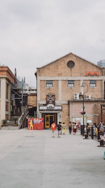 An outdoor space featuring a large brick building with industrial architectural elements. There are several people walking in the open area, creating a sense of casual movement. The front of the building includes a colorful entrance and a poster on the left side showcasing a musical or theatrical event. The surrounding area appears to be an urban setting, with stairs leading to a higher level on the left. Overcast skies contribute to a muted lighting effect.