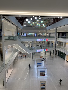 A spacious modern shopping mall with multiple levels and an open atrium. Few people are walking around on the shiny, marble-like floor. The design includes escalators, glass railings, and contemporary art installations. Bright, colorful digital displays adorn the walls.