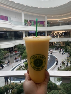 A hand is holding a large cup of orange bubble tea with a green straw in the foreground. The cup features a logo that reads 'Real Fruit Bubble Tea.' In the background, a spacious and modern mall is visible, with multiple levels, bright lighting, and greenery. People are seen sitting and walking around a central area.
