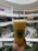 A hand is holding a large cup of orange bubble tea with a green straw in the foreground. The cup features a logo that reads 'Real Fruit Bubble Tea.' In the background, a spacious and modern mall is visible, with multiple levels, bright lighting, and greenery. People are seen sitting and walking around a central area.