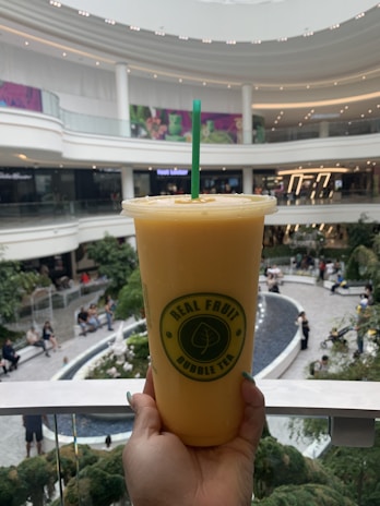 A hand is holding a large cup of orange bubble tea with a green straw in the foreground. The cup features a logo that reads 'Real Fruit Bubble Tea.' In the background, a spacious and modern mall is visible, with multiple levels, bright lighting, and greenery. People are seen sitting and walking around a central area.
