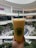 A hand is holding a large cup of orange bubble tea with a green straw in the foreground. The cup features a logo that reads 'Real Fruit Bubble Tea.' In the background, a spacious and modern mall is visible, with multiple levels, bright lighting, and greenery. People are seen sitting and walking around a central area.