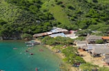 A scenic coastal area with clear turquoise waters and a sandy beach bordered by lush, green hills. The area is dotted with small boats and numerous people enjoying the water and beach activities. Traditional thatched-roof structures and rustic buildings line the edge of the beach, adding a cultural touch to the landscape.