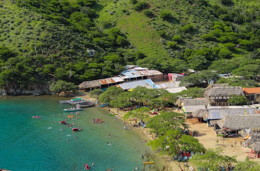 A scenic coastal area with clear turquoise waters and a sandy beach bordered by lush, green hills. The area is dotted with small boats and numerous people enjoying the water and beach activities. Traditional thatched-roof structures and rustic buildings line the edge of the beach, adding a cultural touch to the landscape.