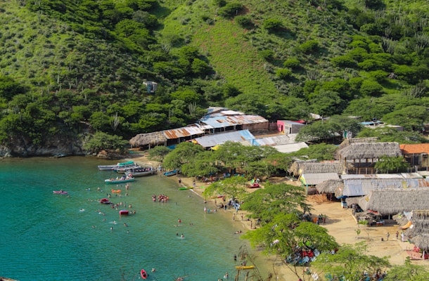 A scenic coastal area with clear turquoise waters and a sandy beach bordered by lush, green hills. The area is dotted with small boats and numerous people enjoying the water and beach activities. Traditional thatched-roof structures and rustic buildings line the edge of the beach, adding a cultural touch to the landscape.