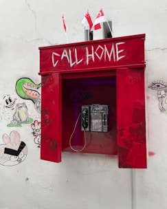 A red, vintage-style telephone booth with 'CALL HOME' written in white, located against a white wall. Beneath the inscription, there is an actual phone with a keypad. The booth is adorned with small Singaporean flags on top. The wall around the booth is decorated with various graffiti, including cartoon-like faces and abstract shapes.