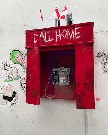 A red, vintage-style telephone booth with 'CALL HOME' written in white, located against a white wall. Beneath the inscription, there is an actual phone with a keypad. The booth is adorned with small Singaporean flags on top. The wall around the booth is decorated with various graffiti, including cartoon-like faces and abstract shapes.