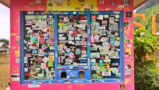 A colorful display covered with an assortment of stickers, each with unique logos, text, and designs. The background is primarily a bright pink structure with blue frames around the sticker-covered panels. Surrounding the structure, there's a glimpse of greenery and other buildings.