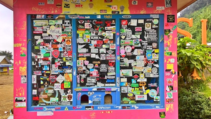 A colorful display covered with an assortment of stickers, each with unique logos, text, and designs. The background is primarily a bright pink structure with blue frames around the sticker-covered panels. Surrounding the structure, there's a glimpse of greenery and other buildings.