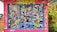 A colorful display covered with an assortment of stickers, each with unique logos, text, and designs. The background is primarily a bright pink structure with blue frames around the sticker-covered panels. Surrounding the structure, there's a glimpse of greenery and other buildings.