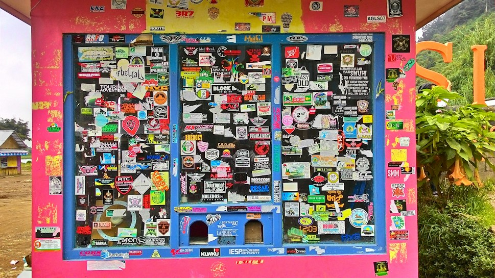 A colorful display covered with an assortment of stickers, each with unique logos, text, and designs. The background is primarily a bright pink structure with blue frames around the sticker-covered panels. Surrounding the structure, there's a glimpse of greenery and other buildings.