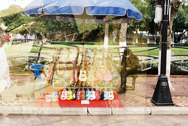 A display of various colorful ukuleles arranged on a red cloth under a blue umbrella in what appears to be a market or park setting. There is a seated figure in the background wearing a hat, and trees and greenery are visible beyond the market setup. The scene seems to have a double-exposure effect, creating a layered and surreal visual.
