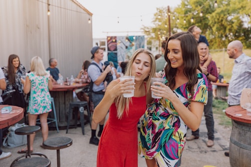A lively outdoor gathering with people socializing around wooden tables. Two women in the foreground are holding drinks and appear to be enjoying themselves. The setting suggests a casual, festive atmosphere with string lights overhead and various groups mingling in the background.