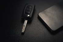 A car key with an extended metal blade is placed next to a modern smartphone on a dark textured surface. The lighting is subtle, creating a sleek and minimalistic aesthetic.