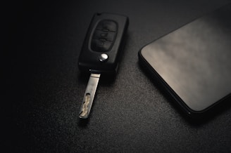 A car key with an extended metal blade is placed next to a modern smartphone on a dark textured surface. The lighting is subtle, creating a sleek and minimalistic aesthetic.