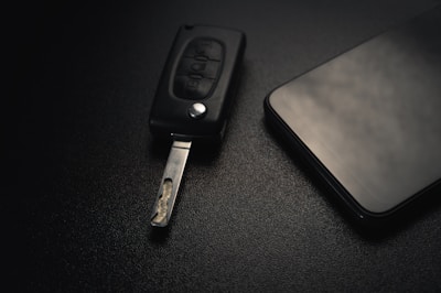 A car key with an extended metal blade is placed next to a modern smartphone on a dark textured surface. The lighting is subtle, creating a sleek and minimalistic aesthetic.
