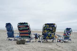 Four beach chairs are arranged on the sandy shore facing the ocean. The chairs feature various colorful patterns, including stripes and floral designs. The sky is overcast, casting a neutral tone over the scene, and the ocean waves are visible in the background. A large rock is positioned in front of the chairs.