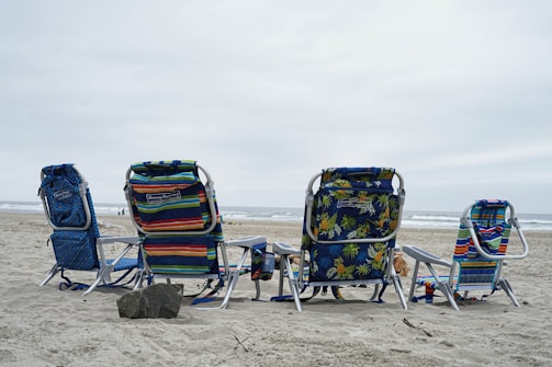 Four beach chairs are arranged on the sandy shore facing the ocean. The chairs feature various colorful patterns, including stripes and floral designs. The sky is overcast, casting a neutral tone over the scene, and the ocean waves are visible in the background. A large rock is positioned in front of the chairs.