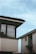 Two modern houses with contrasting architectural designs are positioned under a clear blue sky. One house features large windows with curtains visible, and the rooftops have a dark tiling. The arrangement of the buildings creates sharp lines and angles, lending a contemporary and minimalist feel to the scene.