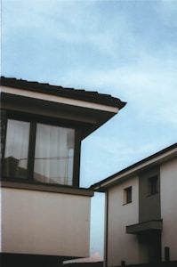 Two modern houses with contrasting architectural designs are positioned under a clear blue sky. One house features large windows with curtains visible, and the rooftops have a dark tiling. The arrangement of the buildings creates sharp lines and angles, lending a contemporary and minimalist feel to the scene.