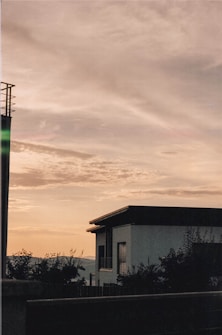 A residential building with a flat roof appears against a backdrop of a serene sky filled with soft clouds. The silhouette of foliage and distant hills can also be seen, suggesting a peaceful suburban or rural setting. The lighting suggests either sunrise or sunset, creating a tranquil atmosphere.